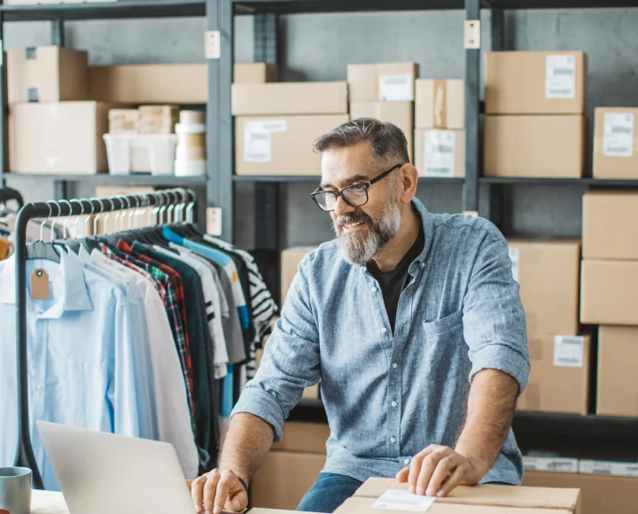Uomo con barba, occhiali e camicia jeans controlla sul laptop l'e-commerce del suo negozio d'abbigliamento